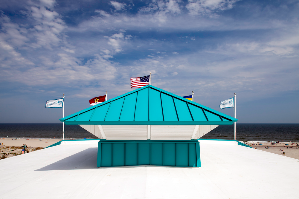 The top of a building looking at a teal metal roof, various flags, and the beach.