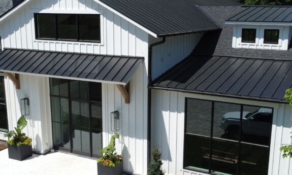 A white board and batten home exterior with a black metal roof surrounded by trees and landscaping.