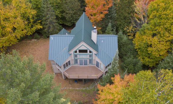 A large home with a blue metal roof surrounded by fall trees in the middle of the woods.